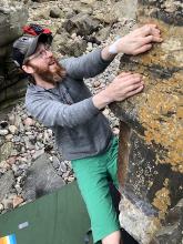 a man with a big red (and grey) beard holding onto a rock while bouldering. He's wearing glasses, a black baseball cap, a grey jumper and green shorts.